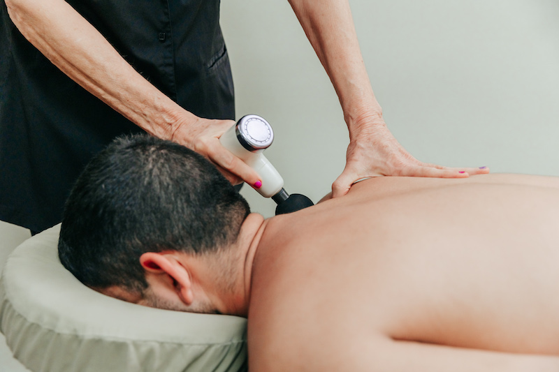 Man lying face-down on a massage table while having deeper tissue massage done to his shoulder using a hand-held device at Anushka's men's salon in West Palm Beach