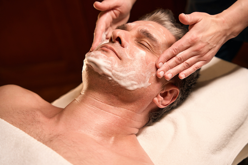 A man lying on a treatment table getting his facial skin cleansed and massaged at Anushka's men's salon in West Palm Beach